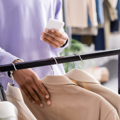 Cropped view of african american showroom owner with smartphone taking jacket from hanging rack